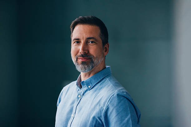 A mature man dressed in a light blue shirt stands confidently, exuding charm and professionalism, with a serene and softly-lit background complementing the composed atmosphere. Perfect for themes of confidence and modern professionals.